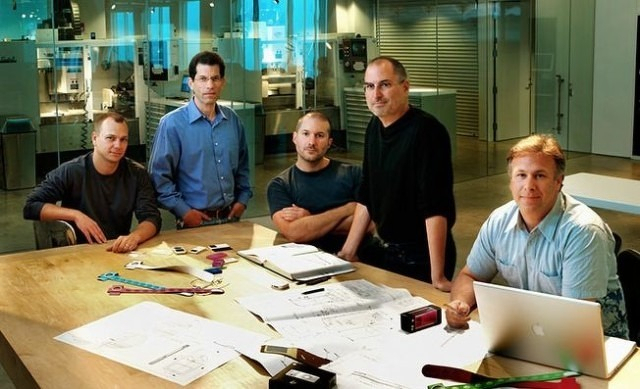 Auto-generated description: Five men are gathered around a table with blueprints and electronic devices in a modern office setting.