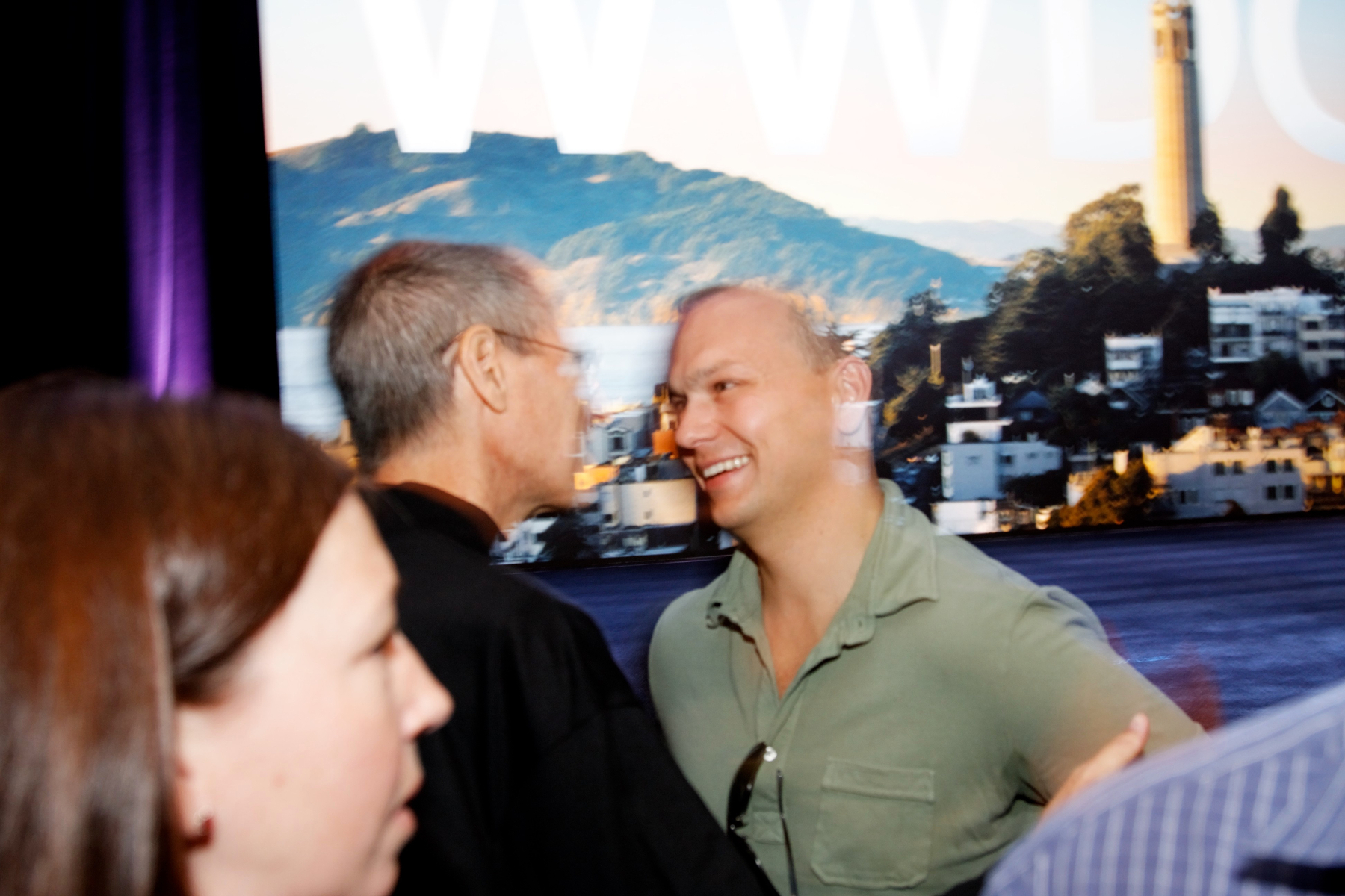 Auto-generated description: Two people are talking and smiling at an event, set against a backdrop featuring a cityscape with hills.