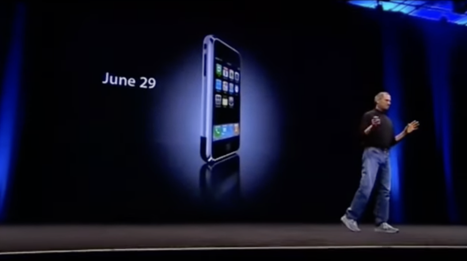 Auto-generated description: A person stands on stage presenting a screen displaying a smartphone and the date June 29.