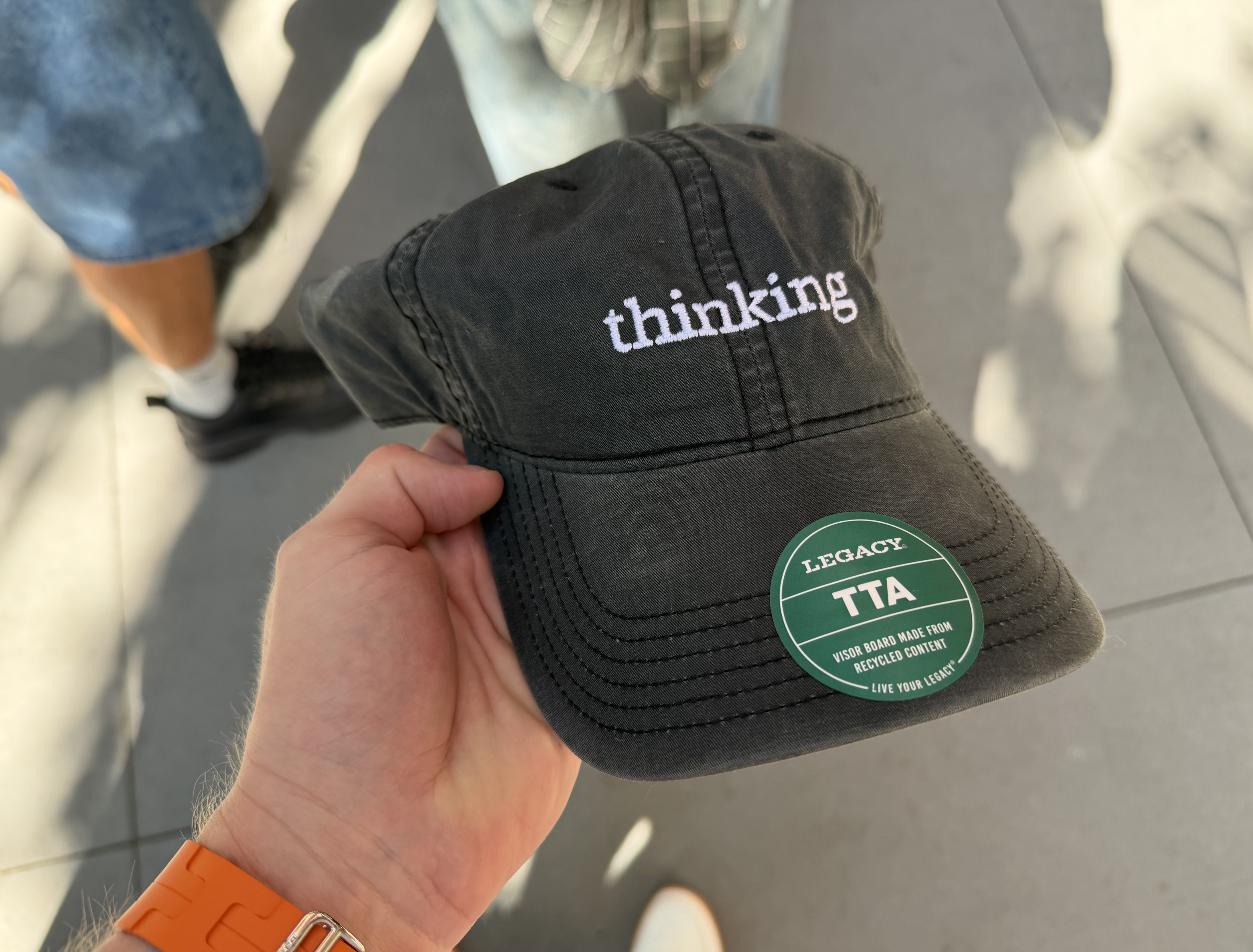 Auto-generated description: A person is holding a black cap with the word thinking embroidered on it, surrounded by people wearing casual attire.