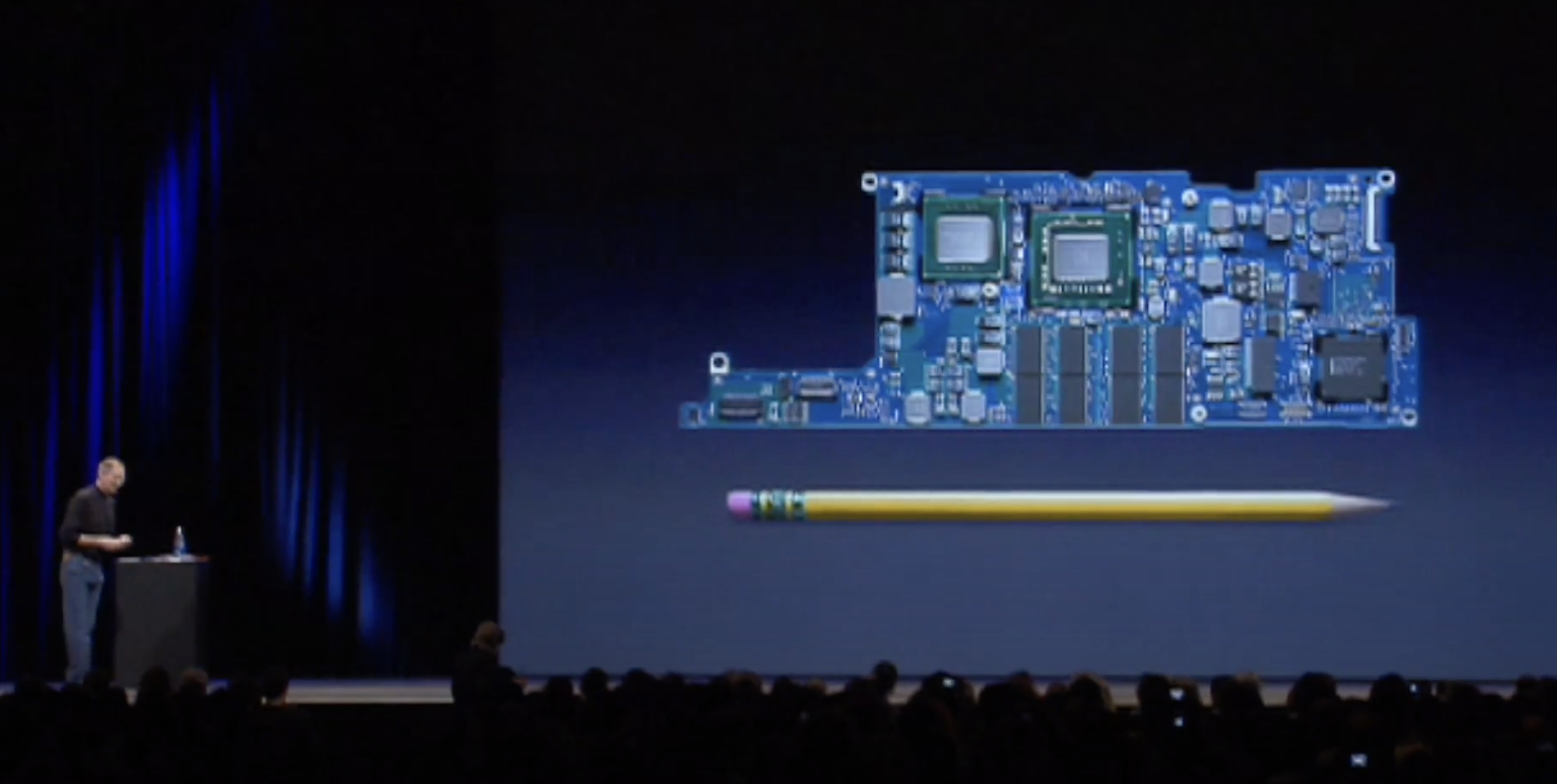 Auto-generated description: A person on stage presents a large image of a computer motherboard compared in size to a pencil.
