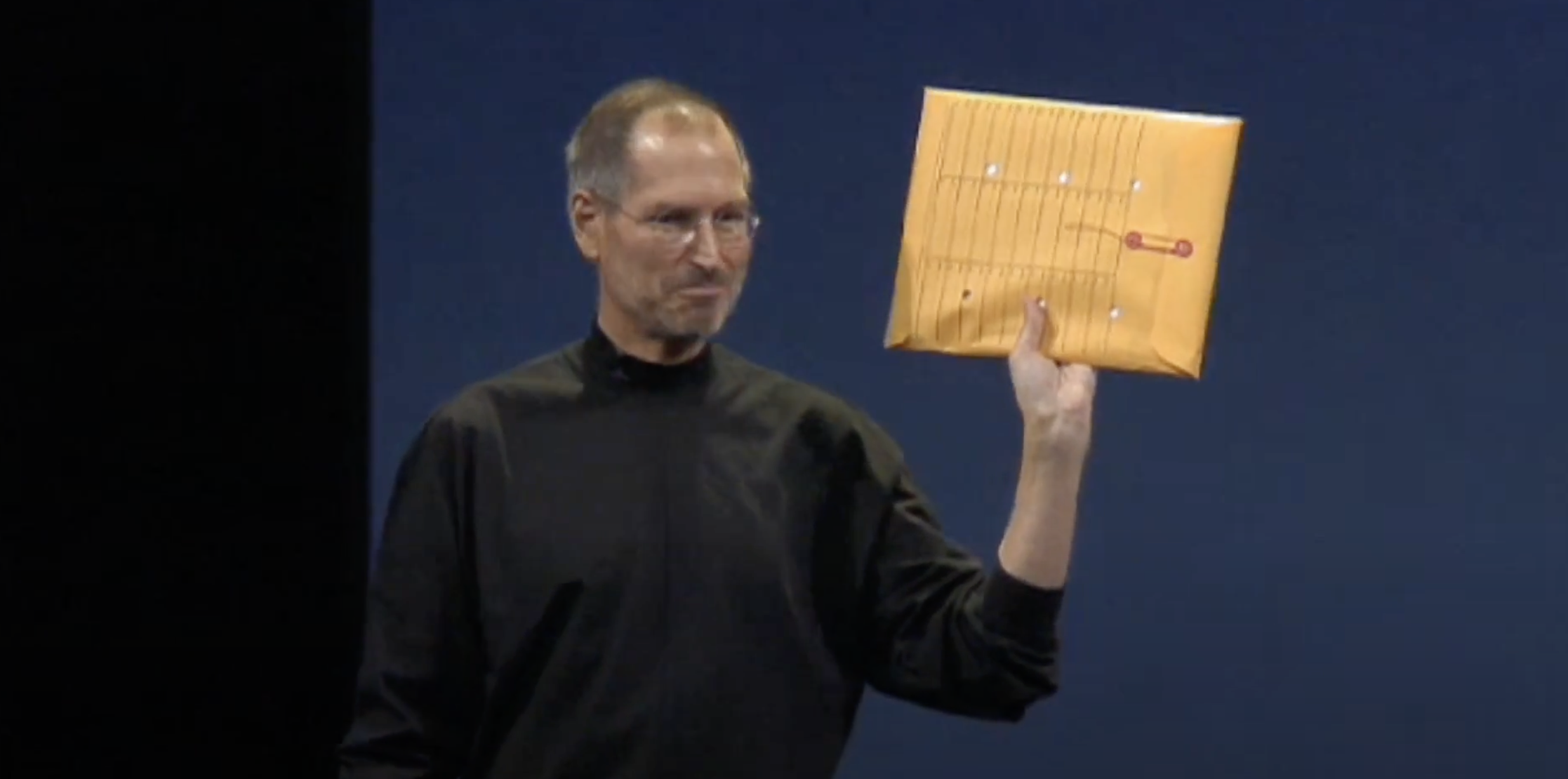 Auto-generated description: A man in a black turtleneck is holding a sealed manila envelope on stage.