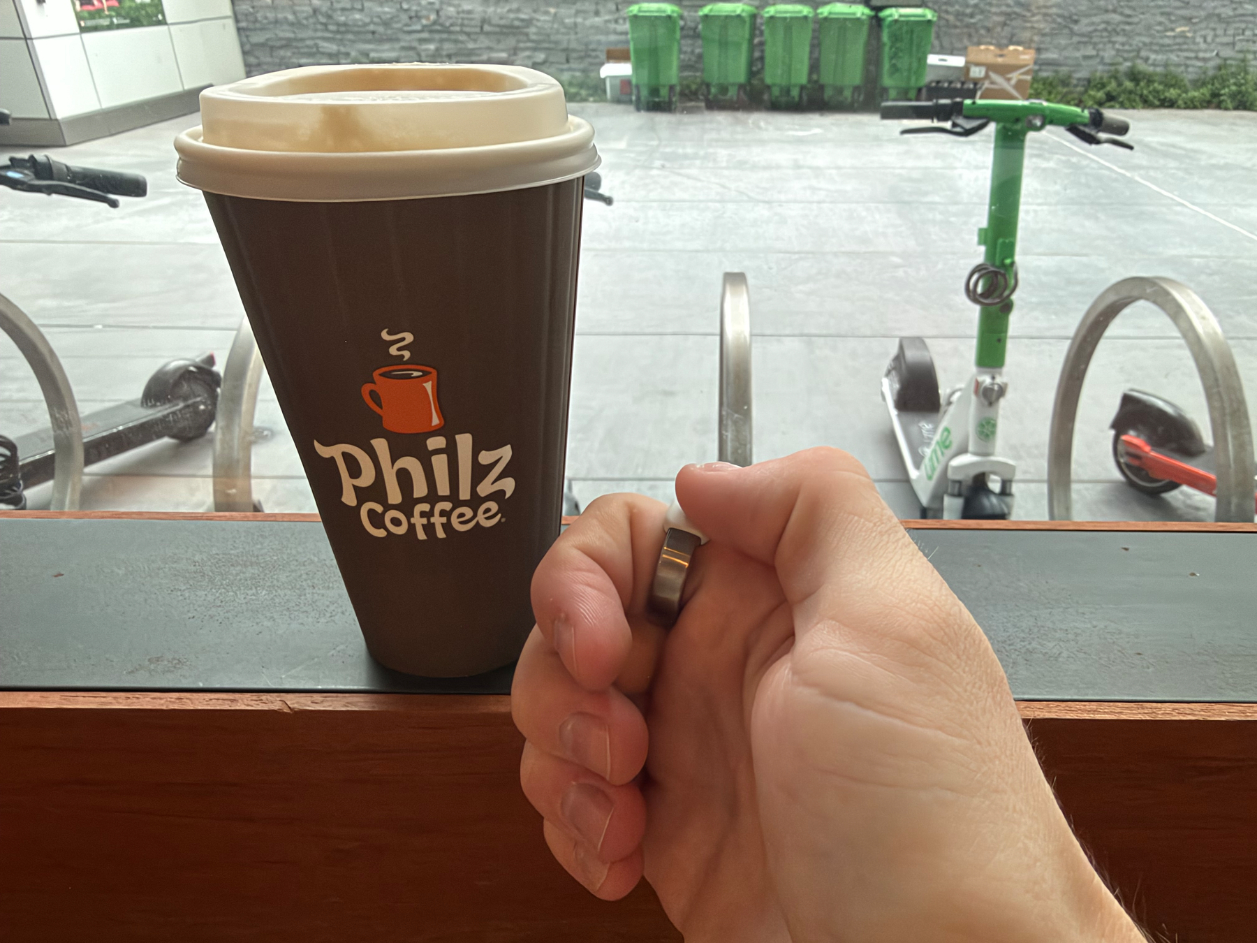A hand is positioned near a Philz Coffee cup on a table with scooters and green trash bins visible outside through a window.